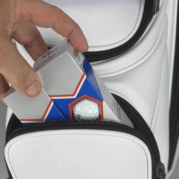 Hand opening a box of golf balls with a white golf bag in the background