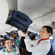 Passenger holding a blue suitcase above their head in an airplane cabin.